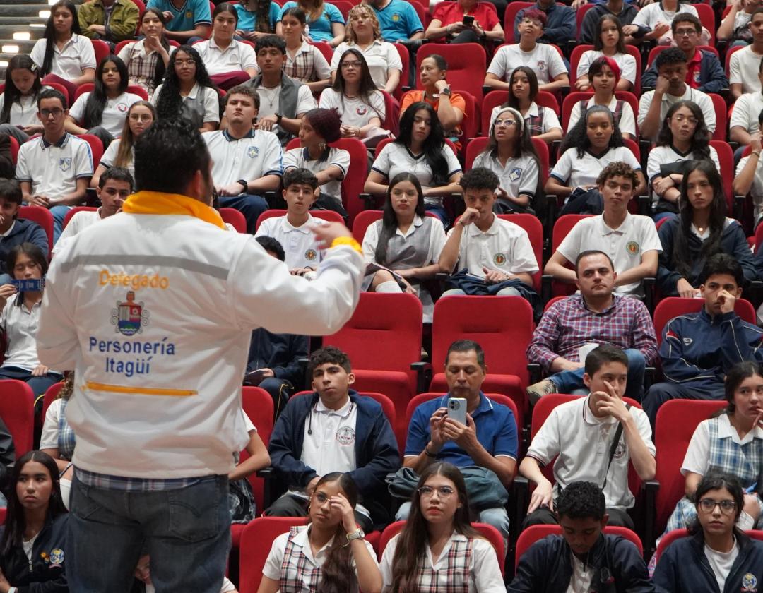 La imagen muestra un auditorio lleno de estudiantes con uniforme escolar, sentados en sillas rojas, mientras escuchan atentamente a un orador. En primer plano, de espaldas a la cámara, hay un hombre con una chaqueta blanca y amarilla que dice 